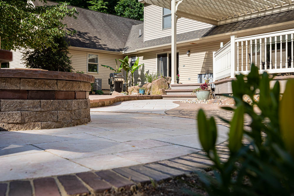 Paver patio, curved stone wall, and planted beds beside a deck and pergola
