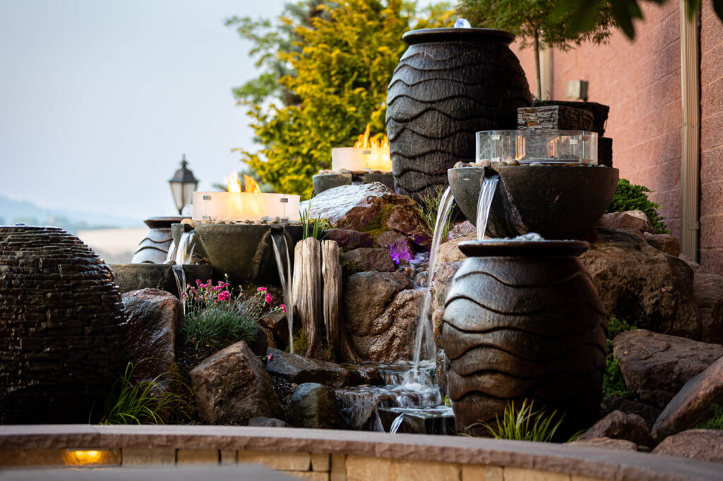 Water cascading from dark urn fountains over rocks beside glass-enclosed fire bowls